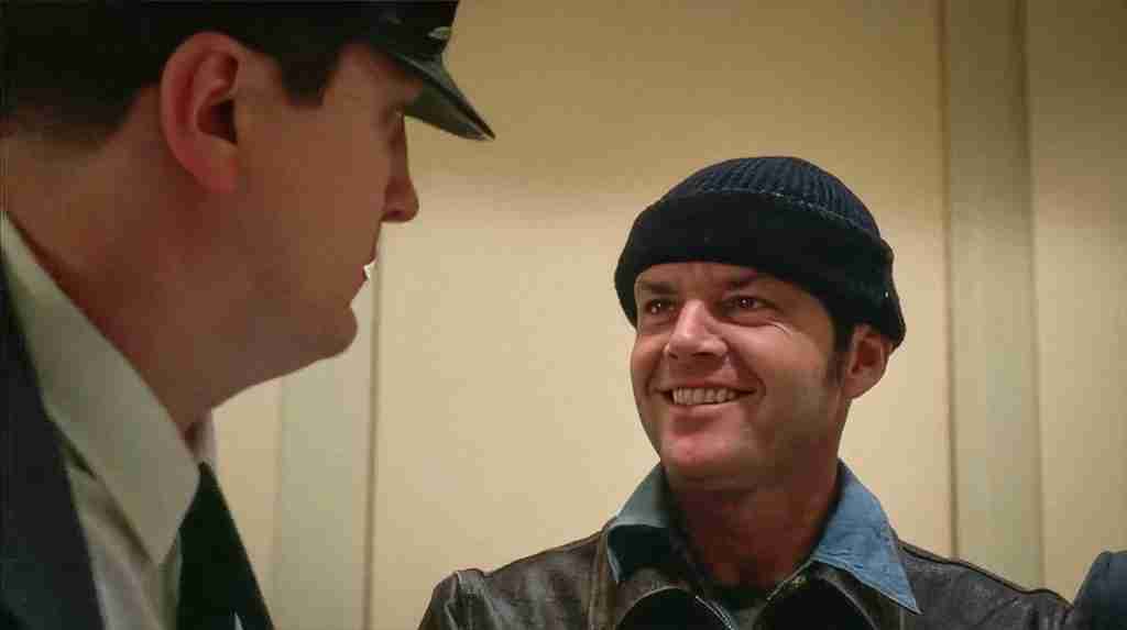 Jack Nicholson as R.P. McMurphy grinning and laughing during his arrival at the mental hospital, escorted by a police officer in One Flew Over the Cuckoo's Nest (1975), marking the inciting incident and disruption of the ward
