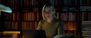 Woman with blonde hair and glasses typing on a laptop at a desk surrounded by tall bookshelves filled with books, illuminated by a green desk lamp in a dimly lit room.