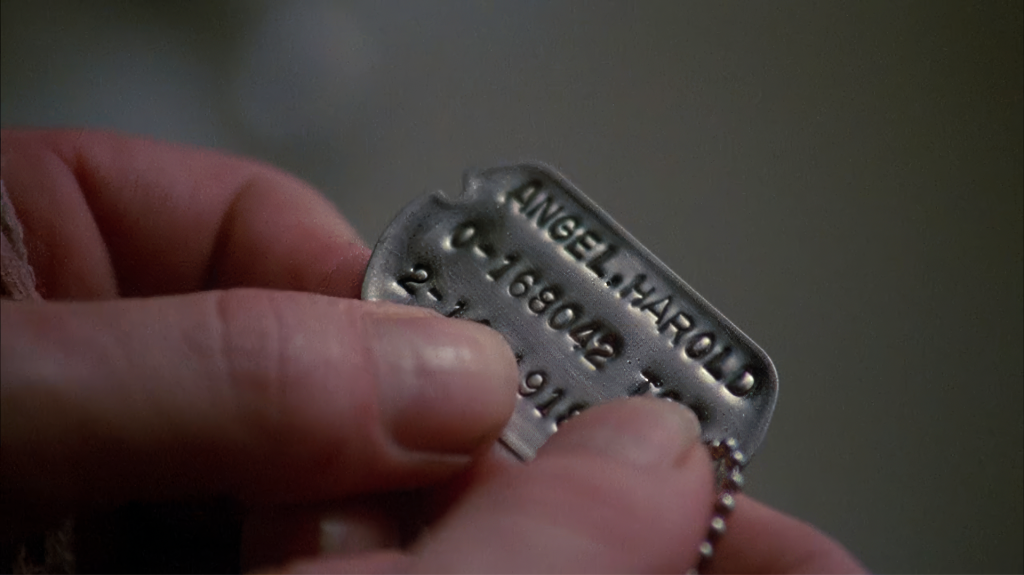 Angel Heart dog tags — close-up of Harry Angel's fingers reading the embossed name ANGEL, HAROLD on military dog tags found in Margaret Krusemark's apartment, the moment the detective discovers he is the missing person