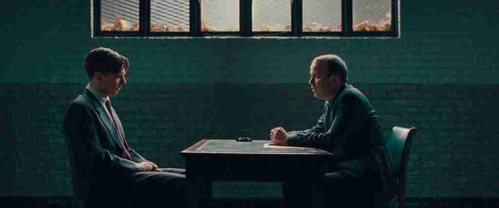 Detective Nock sits across from a somber Alan Turing in a dark interrogation room, setting up the frame story of concealment.
