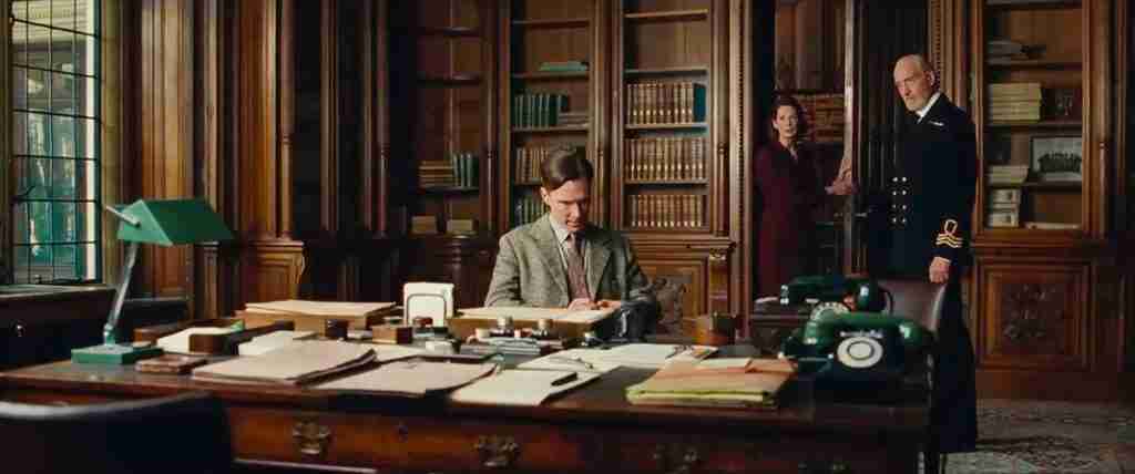 Alan Turing seated in Commander Denniston's office, looking focused as Denniston glares after Alan mentions the Enigma machine.