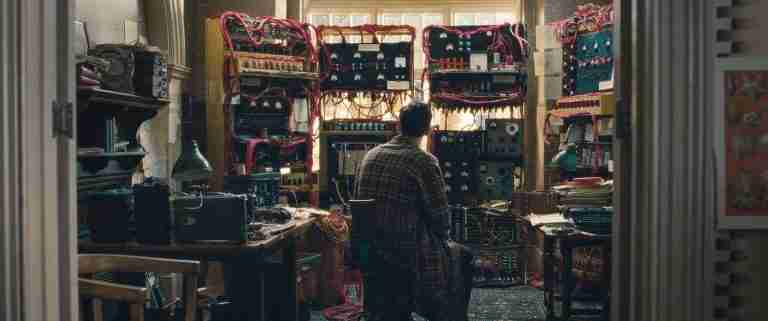 Alan Turing alone, devastated, wearing a dressing gown, staring at his complex codebreaking machine (Christopher) in his home after his arrest.