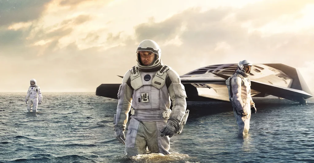 Interstellar analysis still: three astronauts in white pressure suits wade through knee-deep ocean water, a wedge-shaped Ranger spacecraft grounded behind them under a pale amber sky.