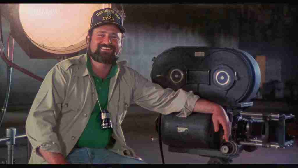Rob Reiner as Marty DiBergi smiling next to a film camera and spotlight, wearing a USS Odonal Sea cap, in the mockumentary This Is Spinal Tap.
