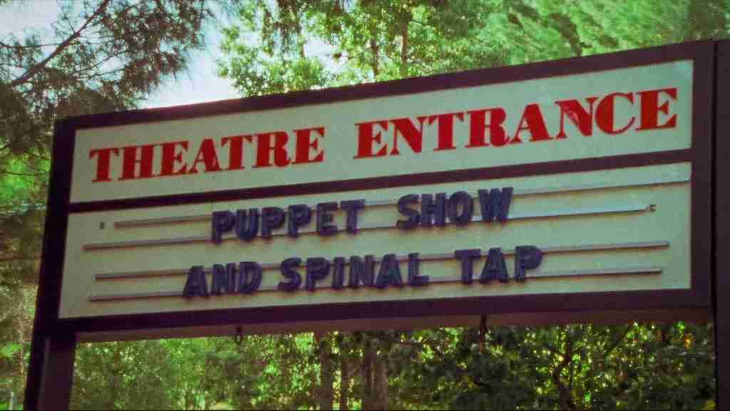 Outdoor theater marquee sign reading "THEATRE ENTRANCE" at the top, with changeable letters below stating "PUPPET SHOW" on one line and "AND SPINAL TAP" on the next, surrounded by trees, in a scene from Rob Reiner's mockumentary This Is Spinal Tap.