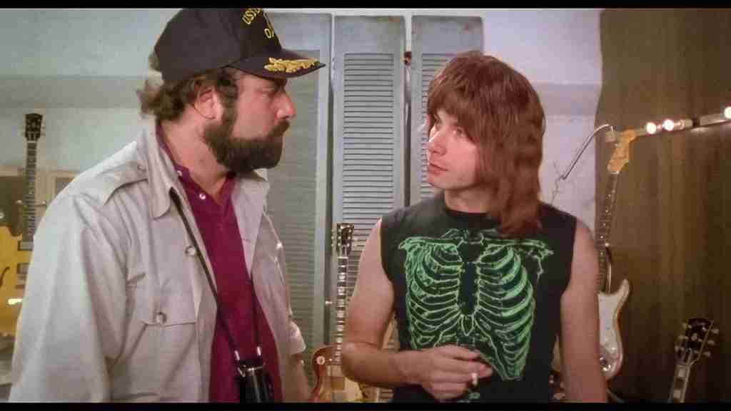 Rob Reiner as Marty DiBergi (left, wearing cap and jacket) talking to Christopher Guest as Nigel Tufnel (right, with long hair and skeleton shirt) in front of guitars and amplifiers during the "These go to eleven" scene in Rob Reiner's mockumentary This Is Spinal Tap.