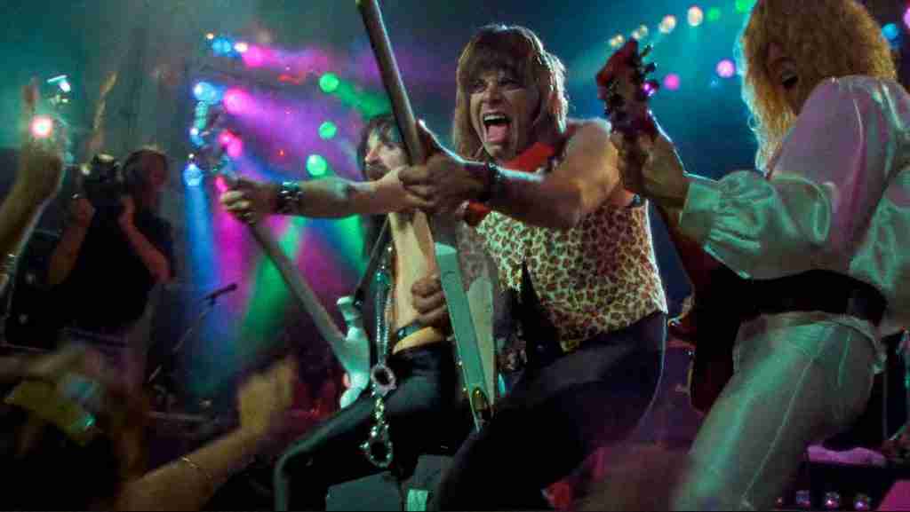 Harry Shearer as Derek Smalls (left), Christopher Guest as Nigel Tufnel (center), and Michael McKean as David St. Hubbins (right) performing together at the front of the stage, striking a unified rock pose under colorful lights as they conclude "Tonight I'm Gonna Rock You Tonight" in Rob Reiner's mockumentary This Is Spinal Tap.