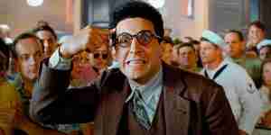 John Turturro as Barton Fink, in glasses and a suit, points frantically at his head in a crowded room, symbolizing the intense creative conflict in the screenwriter vs producer vision debate.