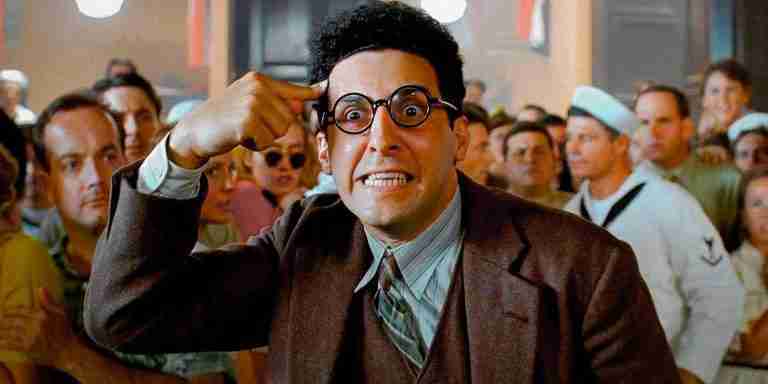 John Turturro as Barton Fink, in glasses and a suit, points frantically at his head in a crowded room, symbolizing the intense creative conflict in the screenwriter vs producer vision debate.