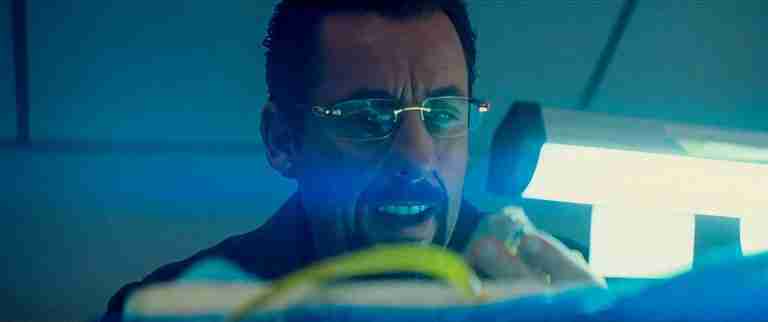 Adam Sandler as Howard Ratner in Uncut Gems, bathed in blue light while obsessively inspecting the black opal.