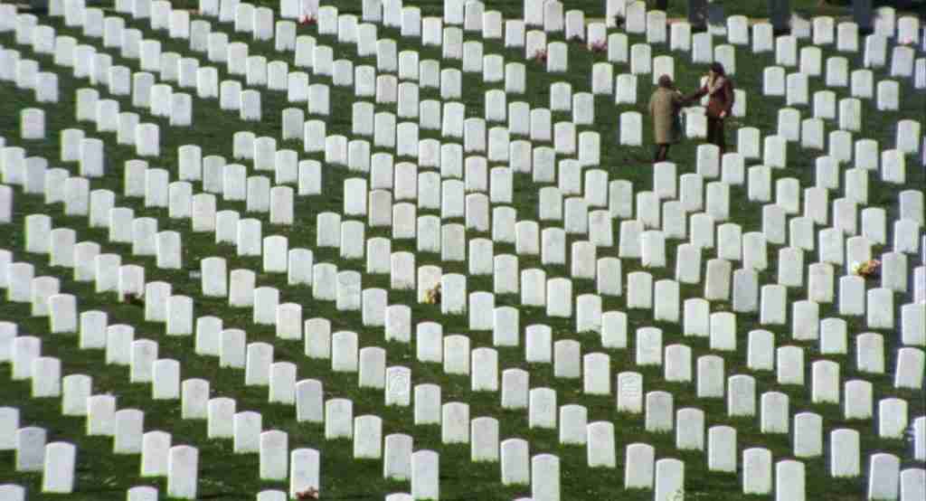 A wide cinematic shot of Harold and Maude standing among thousands of identical white military gravestones, illustrating the theme of conformity.