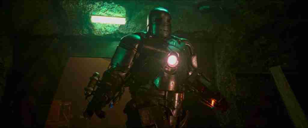 Tony Stark in the bulky Mark I Iron Man armor emerging from the dark cave workshop.