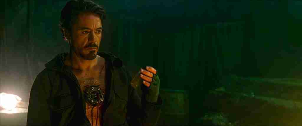 Tony Stark looking at shrapnel fragments in his hand while Yinsen explains the electromagnet.