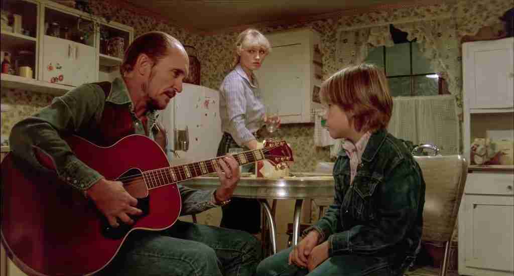 Robert Duvall as Mac Sledge teaching guitar chords to Sonny in a kitchen while Rosa Lee watches in the background.