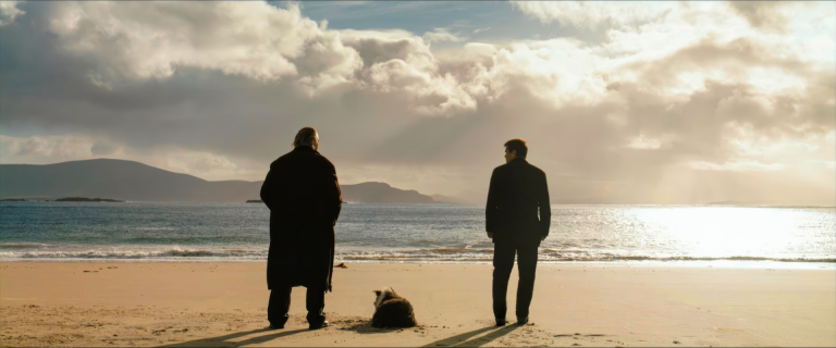 Colm and Pádraic stand apart on the beach with Colm's dog between them in the final scene of The Banshees of Inisherin, the Irish mainland visible across the water