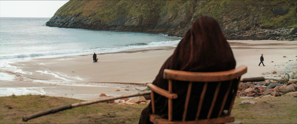Hooded figure of Mrs. McCormick sits watching from the cliff as Colm and Pádraic part on the beach below in The Banshees of Inisherin ending