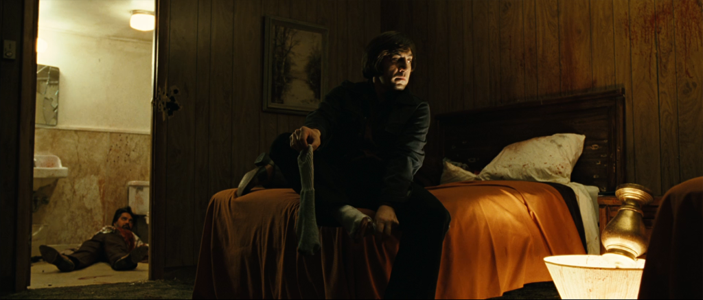 Anton Chigurh sits on a motel bed peeling off blood-soaked socks after killing three Mexican gunmen at Moss's Del Rio motel, with a body visible through the bathroom doorway in No Country for Old Men.