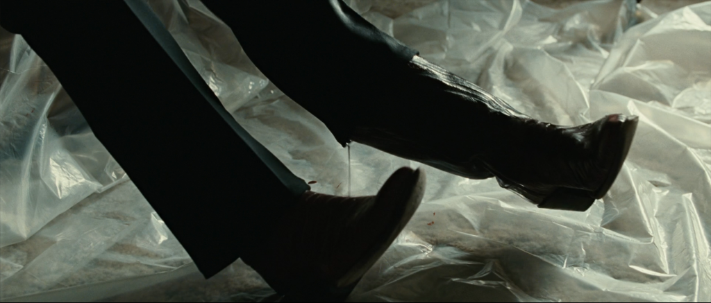 Anton Chigurh sits on a motel bed, using his feet to pry off his boots over a plastic tarp, with his own blood draining from the boot-tops as he prepares to perform self-surgery on the leg wound Moss inflicted at Eagle Pass in No Country for Old Men.