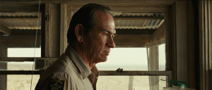 Sheriff Bell war story scene, Tommy Lee Jones as Ed Tom Bell in profile in Ellis's cabin in No Country for Old Men, framed against the West Texas landscape during the moment he tells Ellis that God never came into his life