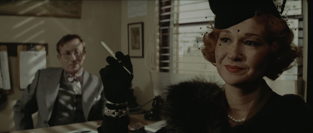 Diane Ladd as Ida Sessions posing as Evelyn Mulwray in Jake Gittes's office, in a black veiled hat, black gloves, fur collar, and pearls, holding a cigarette while a man in a grey suit and bow tie watches from a desk behind her.