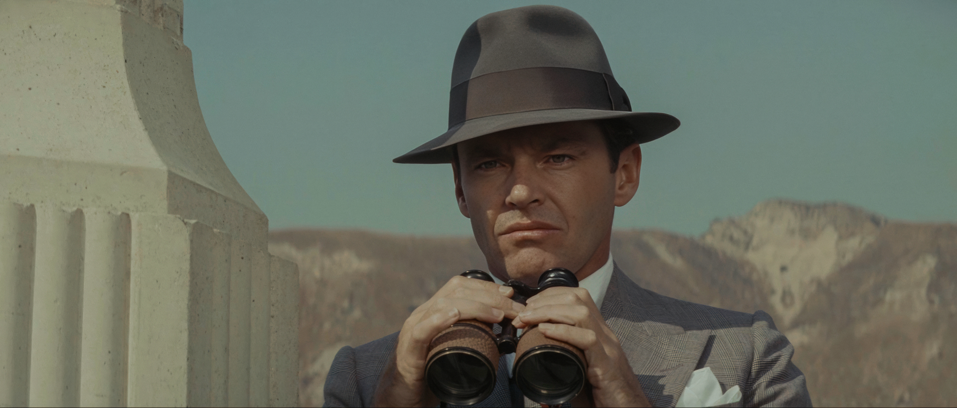 Jack Nicholson as Jake Gittes in a suit and fedora, holding binoculars at his chest, with bare hills behind him above the dry Los Angeles River bed in Chinatown.