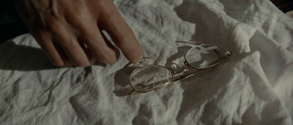 Jake's hand reaching for the broken wire-rimmed bifocals lying on pale rumpled fabric in Chinatown.
