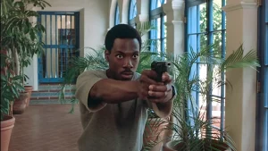 Axel Foley aims a pistol with a two-handed grip in a plant-lined corridor of Maitland's estate in Beverly Hills Cop.