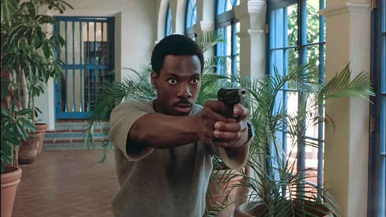 Axel Foley aims a pistol with a two-handed grip in a plant-lined corridor of Maitland's estate in Beverly Hills Cop.