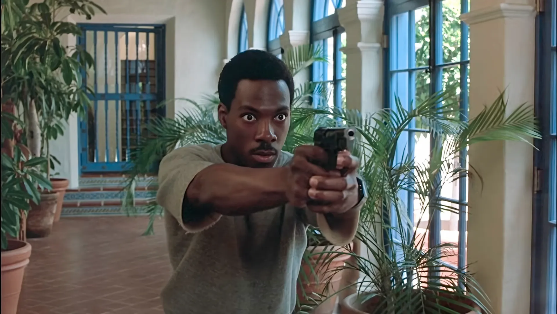 Axel Foley aims a pistol with a two-handed grip in a plant-lined corridor of Maitland's estate in Beverly Hills Cop.