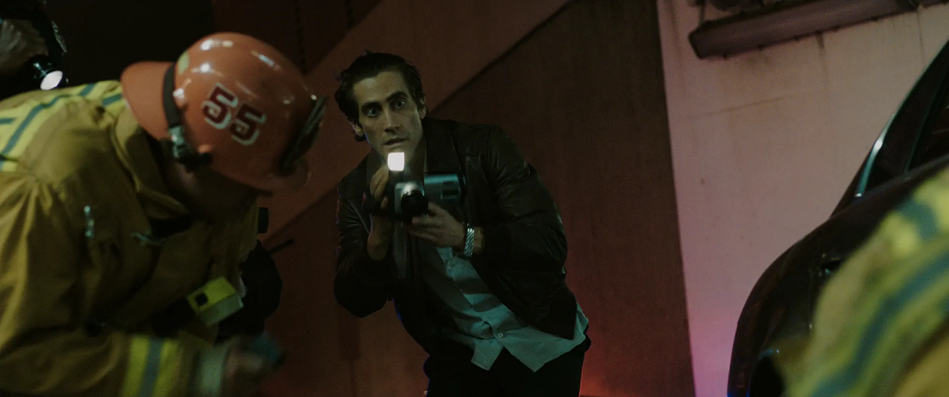 Lou Bloom aims a camcorder light at firefighters working a nighttime scene in Nightcrawler.
