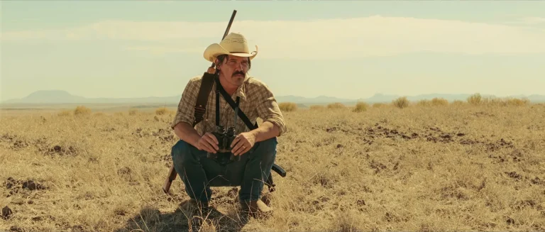 No Country for Old Men book vs movie: Llewelyn Moss (Josh Brolin) crouched in the West Texas grassland, the landscape the film opens on instead of the screenplay's snow