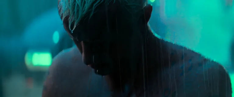 Roy Batty, bare-shouldered and head bowed, rain running down his face and neck, city lights blurred teal behind him