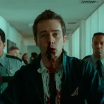 Edward Norton as the narrator, face bloodied and shirt torn, being walked down a bright office corridor by two security guards while coworkers watch, from Fight Club.