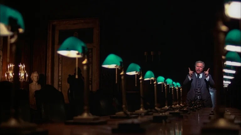 Arthur Jensen (Ned Beatty) standing at the end of a long dark boardroom table lined with green banker's lamps in Network (1976)