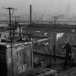Wide shot of a Hoboken rooftop with pigeon coop, Golden Warriors boys, Terry Malloy, and the Manhattan skyline in haze in On the Waterfront